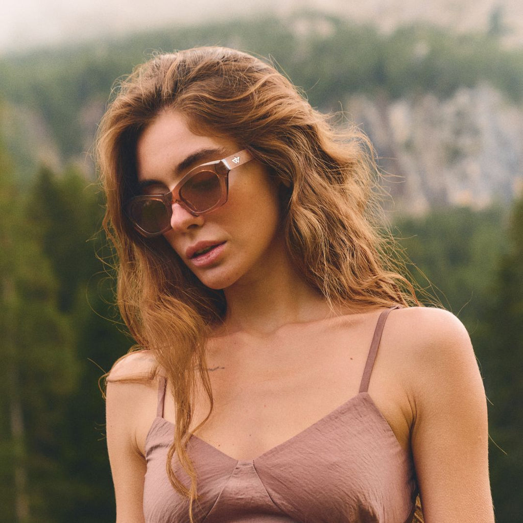 Woman wearing sunglasses and a brown top outdoors with greenery in the background